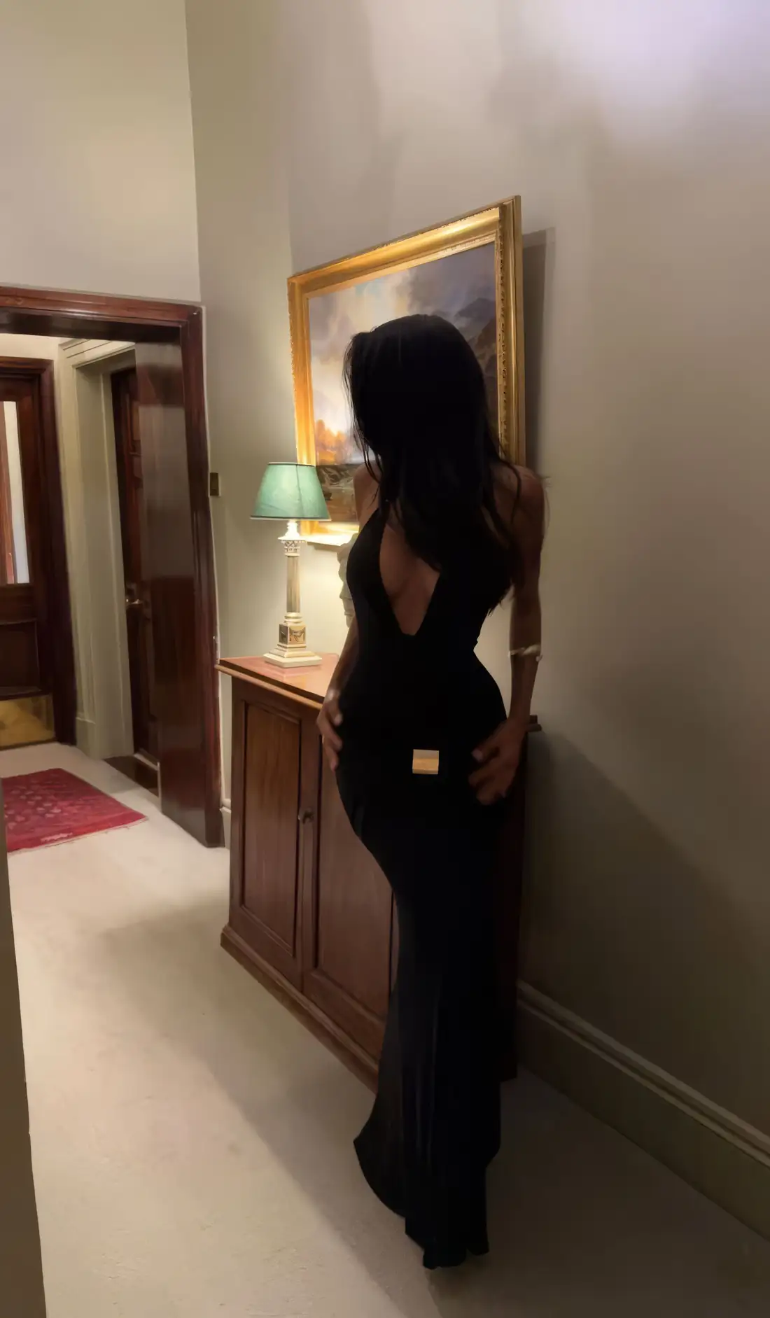 A woman in a black gown in a classic hallway beside a gilt-framed painting